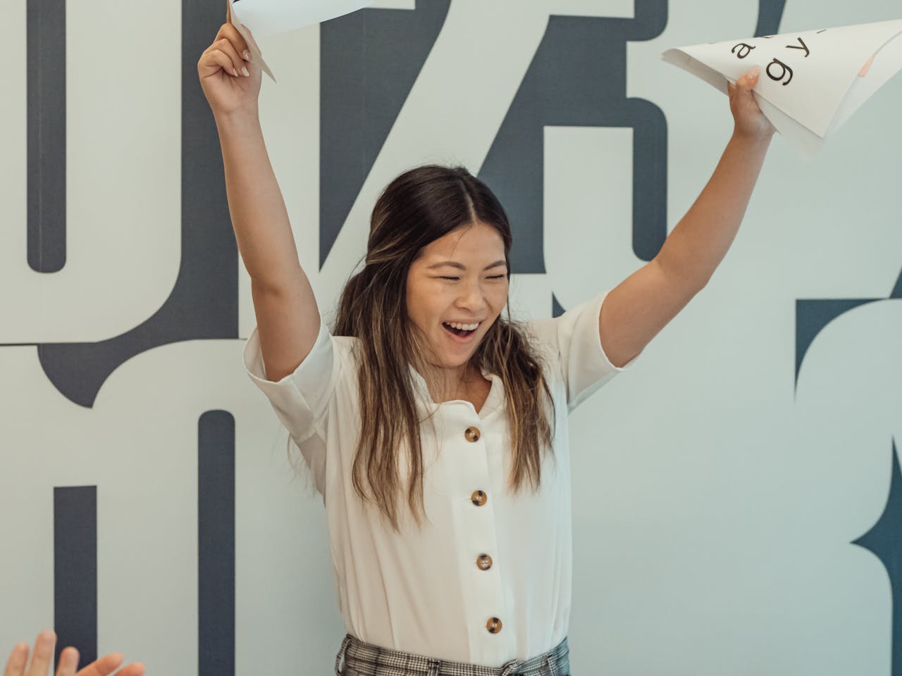 Asian woman in office celebrating success with raised arms holding documents.